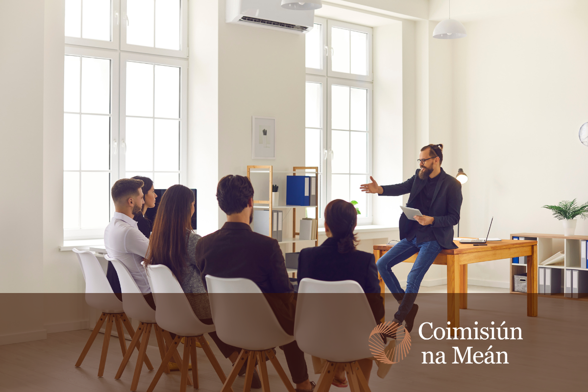 A bearded man dressed in business casual presents his idea to a committee in an office setting